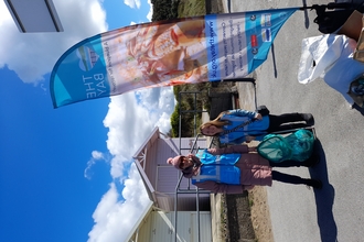 A bright blue sky, under which two women are standing, in front of two beach huts, with litter pickers and a bag of rubbish