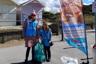 Two women are wearing blue hi vis vests standing with a full bag of rubbish they have collected